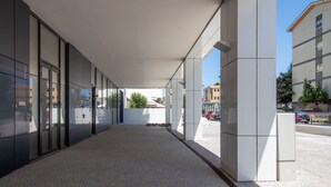 Un pasillo cubierto con puertas de vidrio que conducen a un edificio, cielo despejado y coches estacionados al fondo.