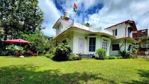 Una casa de dos pisos con fachada blanca, techo rojo e izada de una bandera en la parte superior. Un césped cuidado se extiende al frente, y se observa un paraguas rojo a la izquierda.