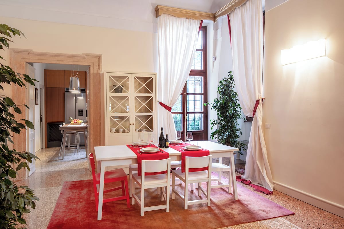 Un comedor con una mesa preparada para cuatro, una cocina al fondo y un ventanal con cortinas.