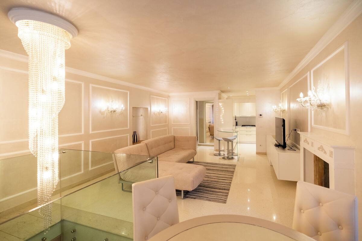 Una sala de estar moderna con una escalera de cristal, un sofá color beige y una araña.