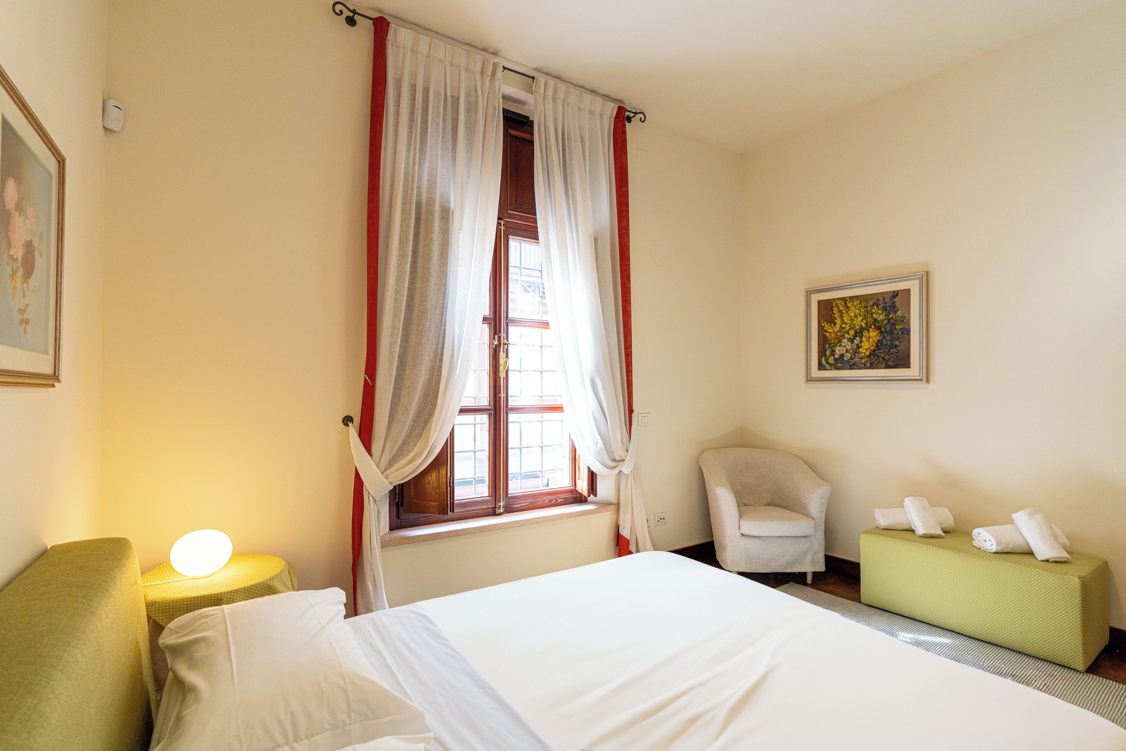 Ein Hotelzimmer mit Bett, Sessel, kleinem Tisch mit Lampe, Vorhängen an einem Fenster und einem Bild an der Wand.