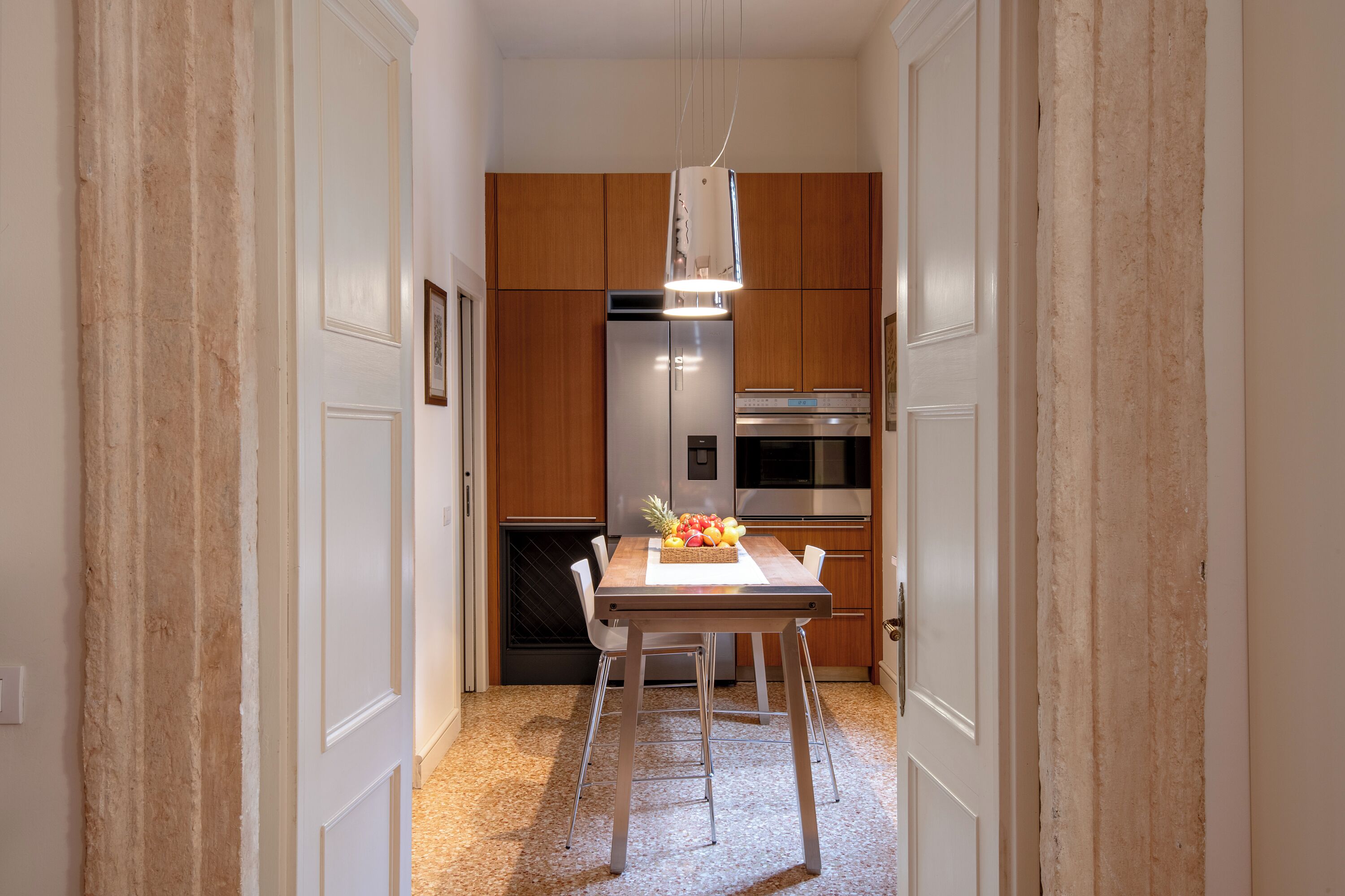 A narrow hallway leading to a kitchen with wooden cabinets, a stainless steel refrigerator, and a dining table with chairs.