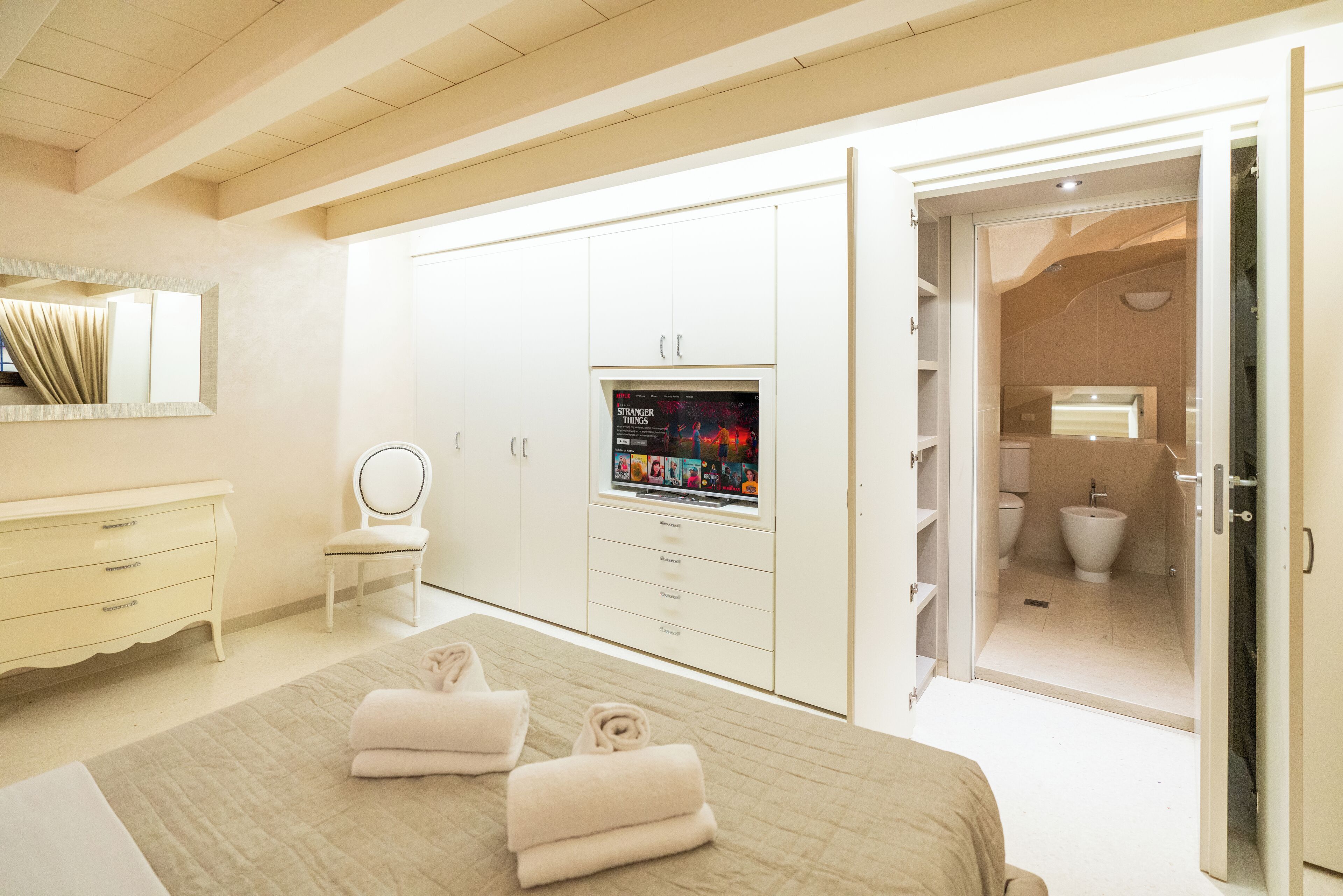 A bedroom with a bed, a TV on a stand, a wardrobe, a bathroom visible through an open door, and a chair.