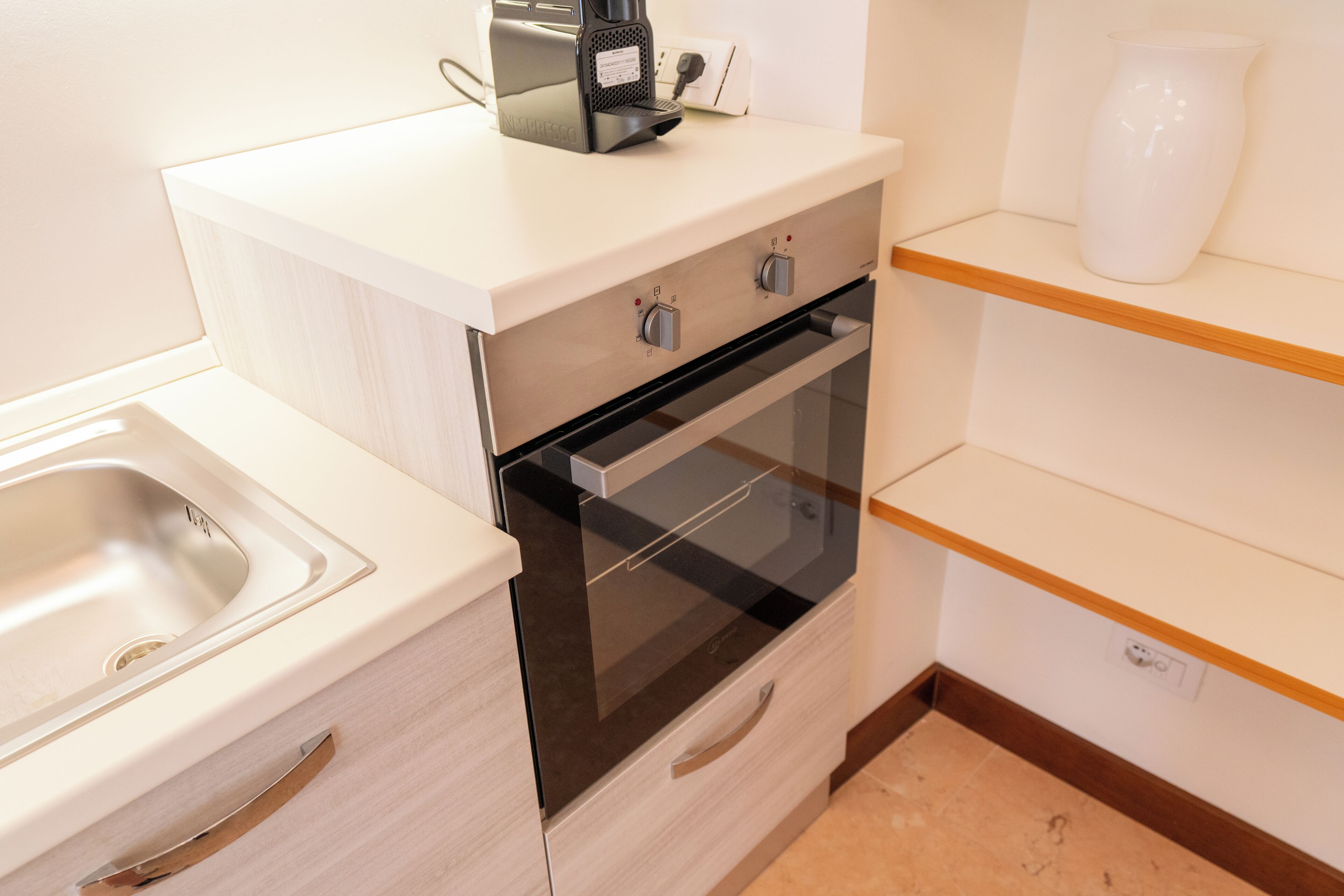 A kitchen with a sink, a coffee maker, a white vase, and wooden shelves.