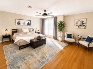 Quarto com cama, mesas de cabeceira, um criado-mudo, ventoinha no teto, planta em vaso, cadeira e janela com cortinas.
