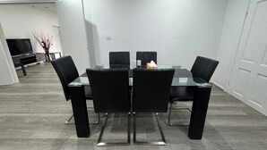 Een moderne eethoek met een glazen tafel en zwarte stoelen, een flatscreen televisie op de achtergrond en een witte deur aan de rechterkant.