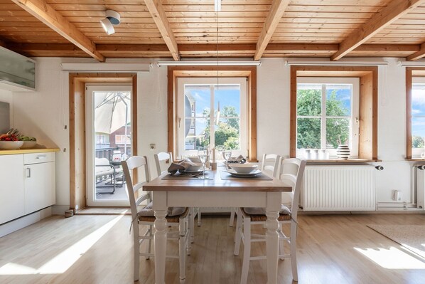 Un espace repas avec une table en bois, des chaises blanches et de grandes fenêtres.