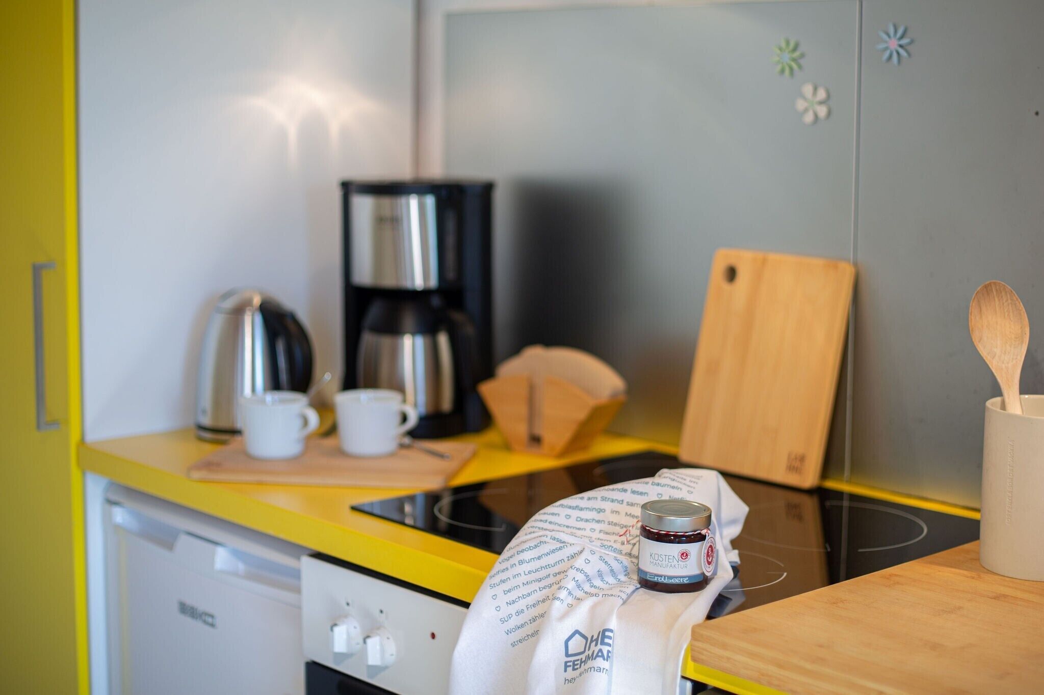 Eine Küche mit gelber Arbeitsplatte, einer Kaffeemaschine, einem Wasserkocher, einem Schneidebrett und einem Marmeladenglas.