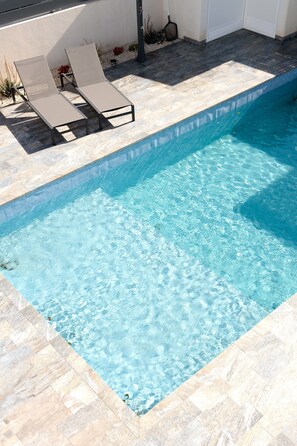 A swimming pool with clear water, surrounded by a tiled deck and two lounge chairs.