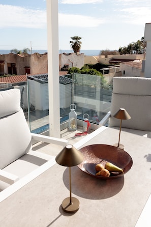 A balcony with a glass railing, white cushions, a table with a bowl of fruit, and a lamp.
