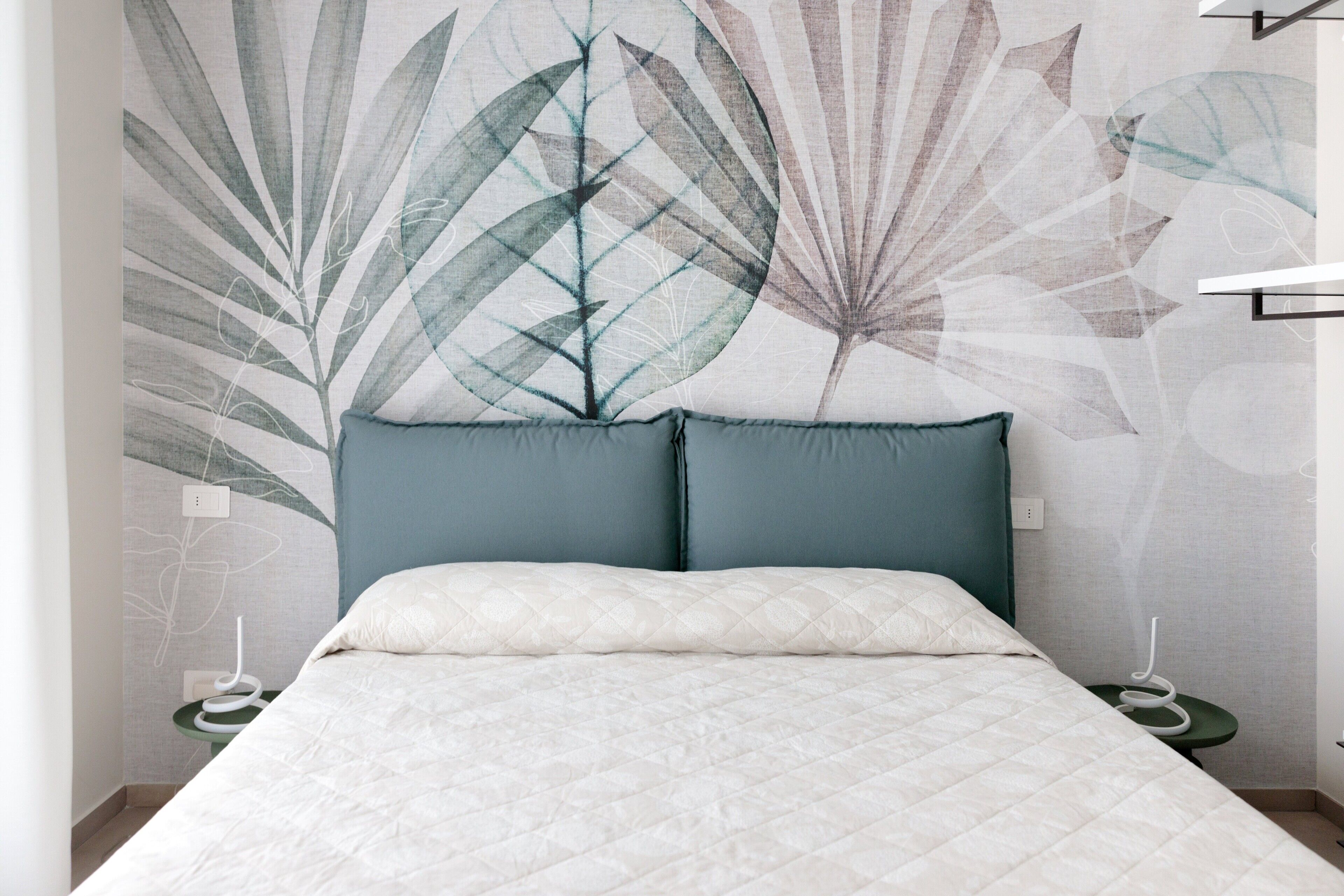 A bed with a quilted white cover, two teal pillows, and a wall with a leaf pattern.