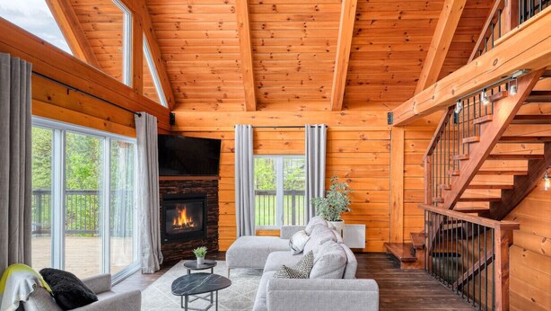 A cozy log cabin interior with a fireplace, comfortable seating, and a staircase. - Henö - Entre lac & forêt (Mandeville)