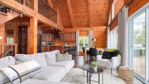 A spacious living area with a sofa, coffee table, and chairs, wooden beams on the ceiling, and large windows offering a view of the outdoors. - Henö - Entre lac & forêt (Mandeville)