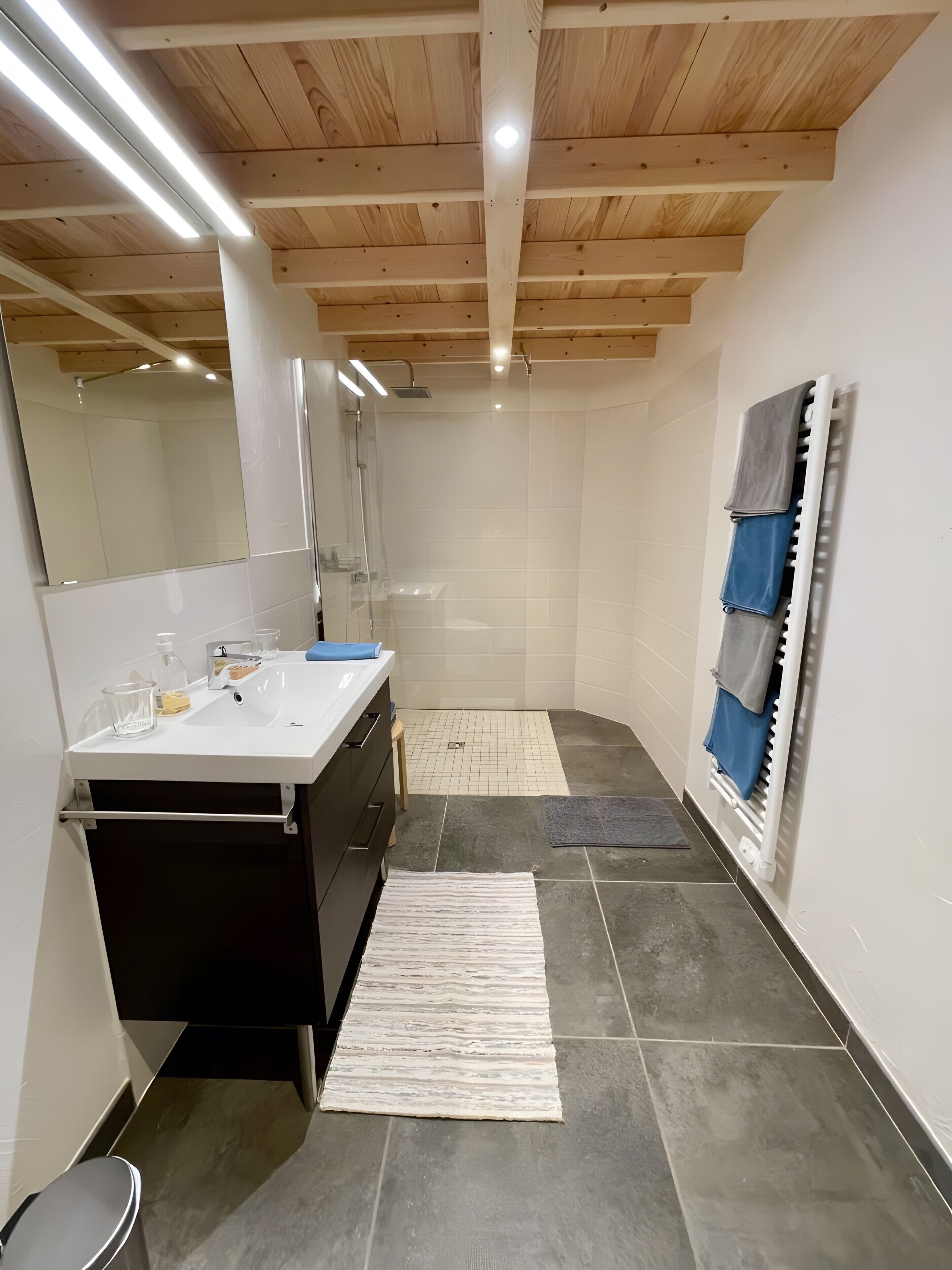 Une salle de bain moderne avec un plafond en bois, un grand miroir, une douche, un lavabo avec des articles de toilette et un porte-serviettes.