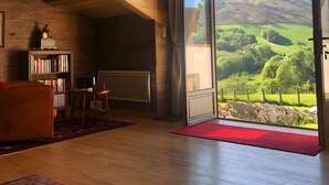 A room with a sloped ceiling, a large window with a view of green hills, a bookshelf, and a red rug. - Guest Room 'Olceiry' with Mountain View, Private Garden and Wi-Fi (Iholdy)