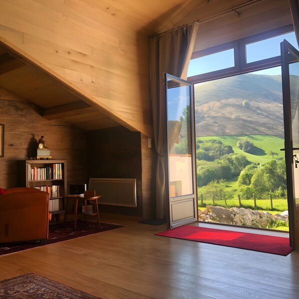 A room with a sloped ceiling, a large window with a view of green hills, a bookshelf, and a red rug.