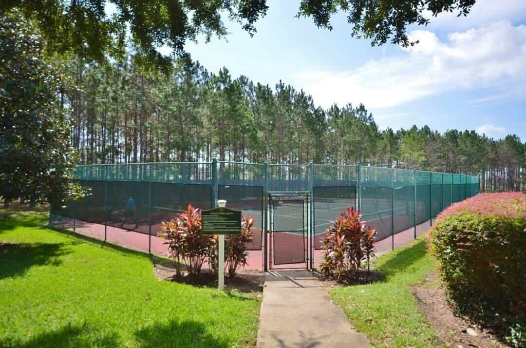 A tennis court enclosed by a green fence, surrounded by trees and a pathway leading to the entrance.