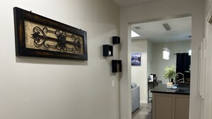 A hallway with a framed artwork, a kitchen area with a sink and countertop, and a gray couch in the background.