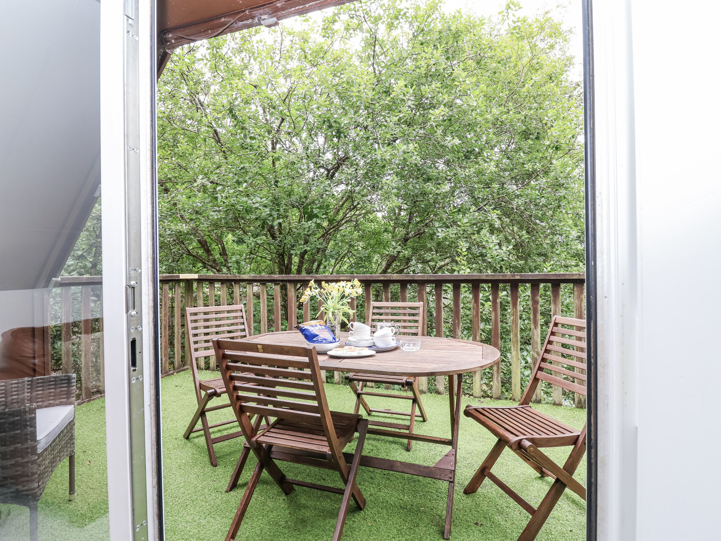 Un patio al aire libre con una mesa y sillas de madera, rodeado por un cerco de madera y abundante follaje.