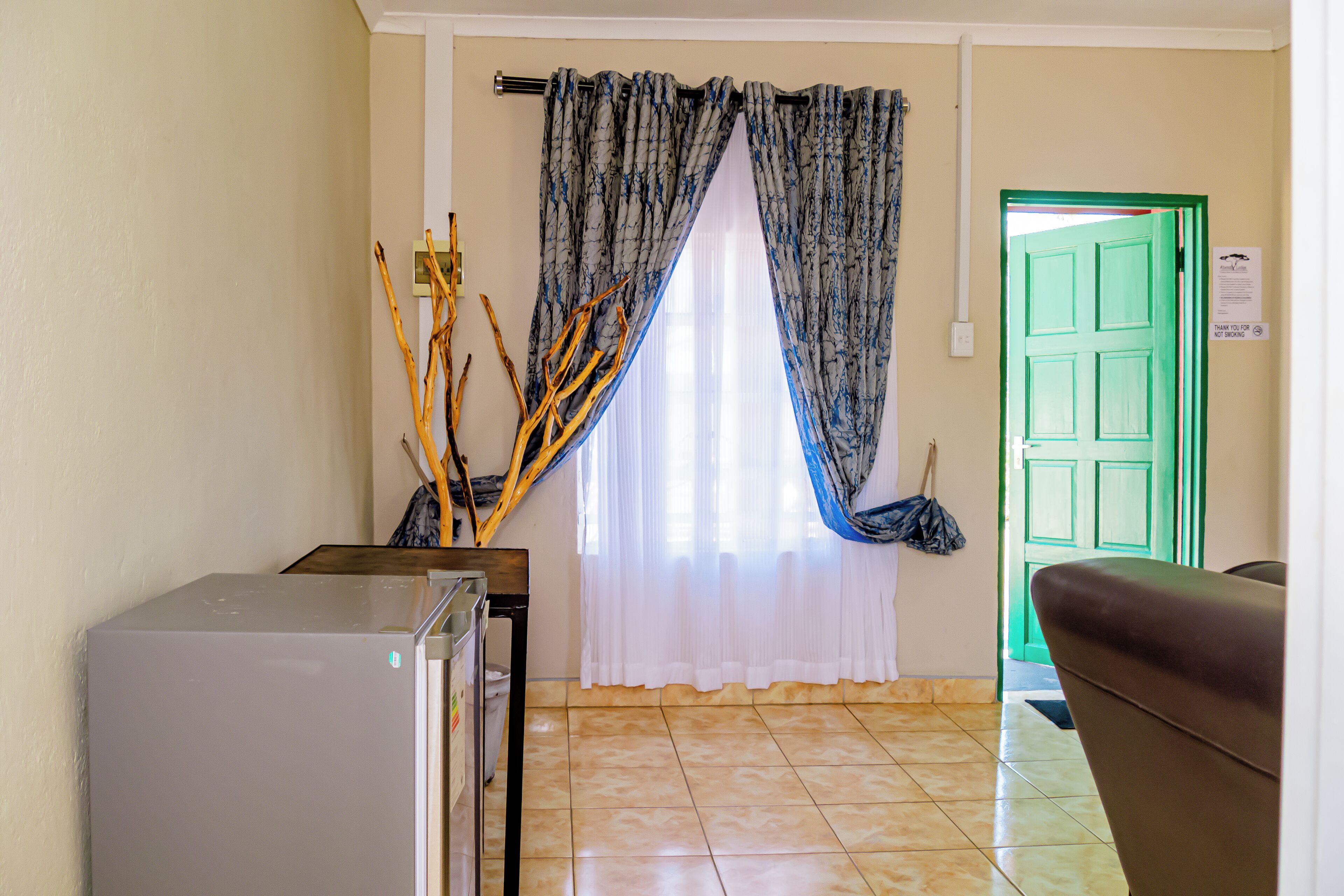 Una habitación con refrigerador, una mesa de madera, una puerta verde y una ventana con cortinas.