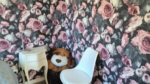 Una habitación con papel tapiz floral, una silla blanca, una mesita de noche de madera y un oso de peluche.