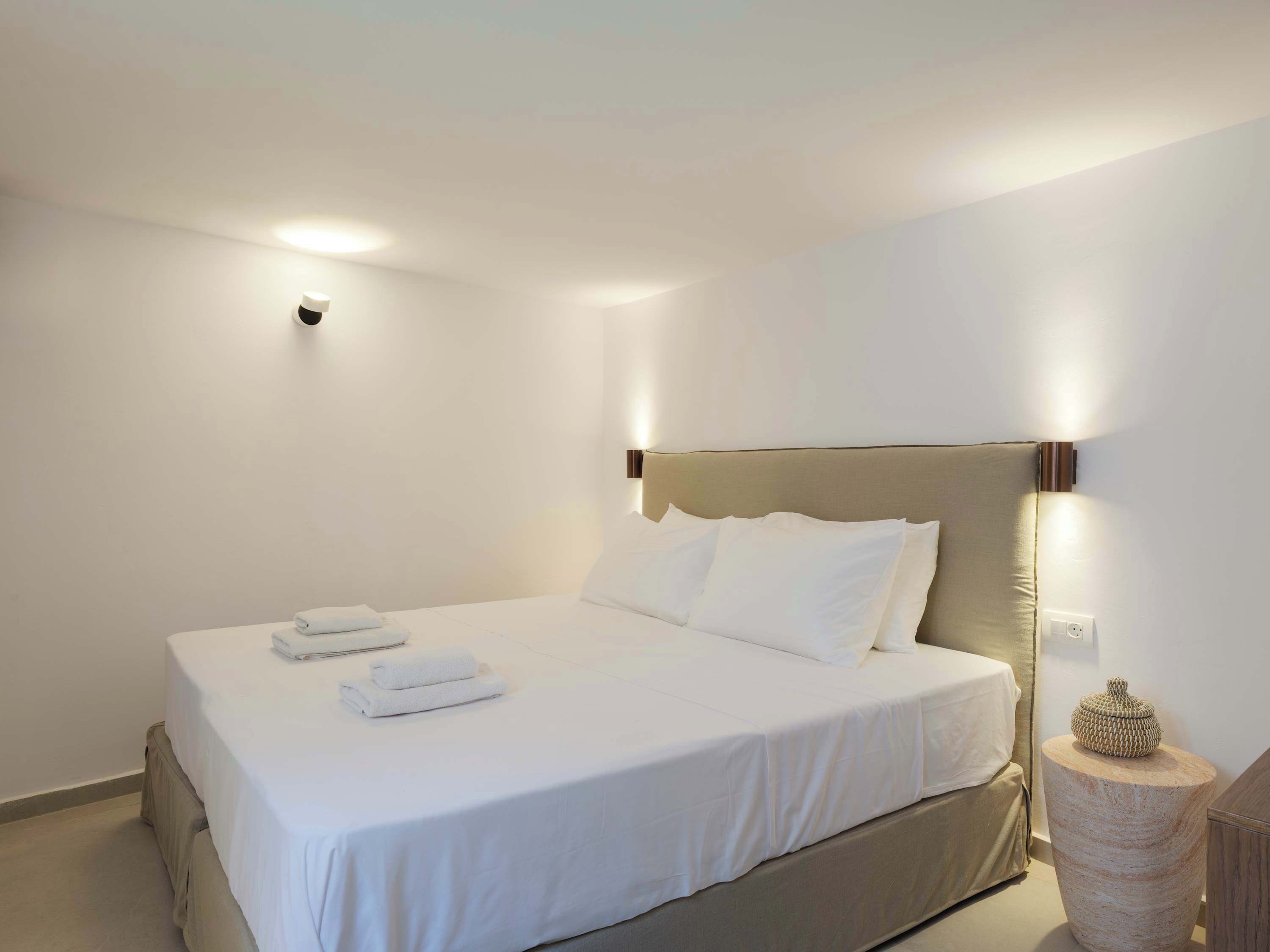 Una cama bien hecha con sábanas blancas y dos toallas dobladas, una mesita de noche redonda de madera con un adorno, y un aplique de pared.