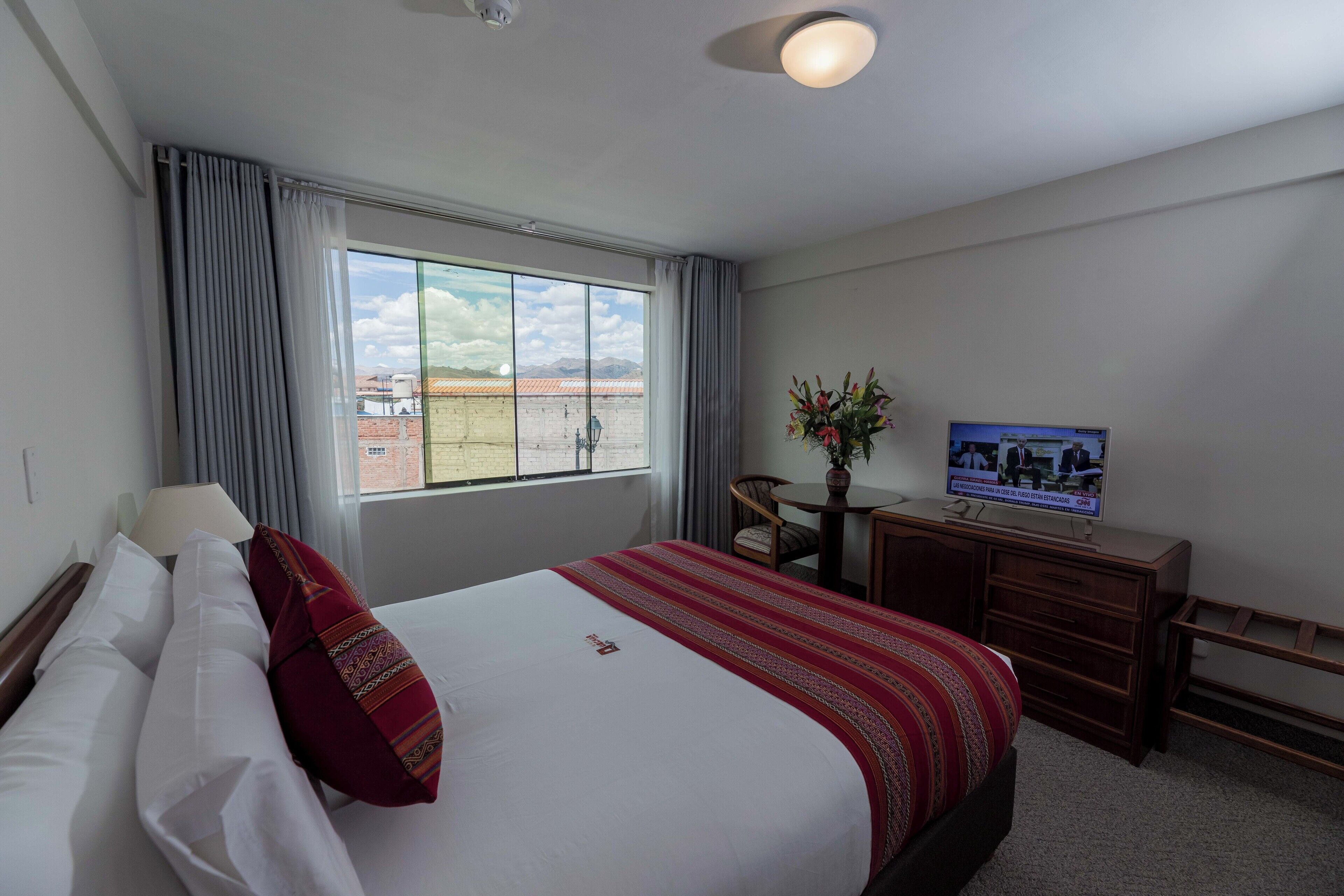 A hotel room with a large bed, a desk with a television, a window with curtains, and a view of buildings outside.