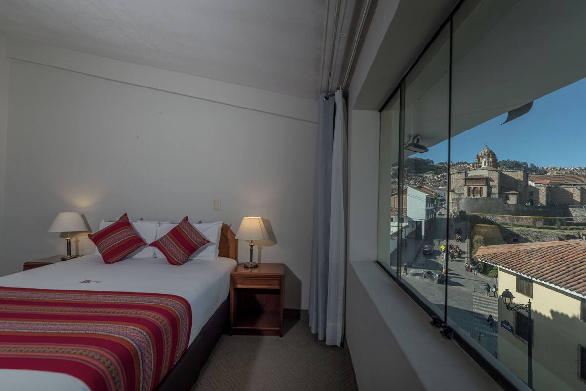 A hotel room with a bed, two bedside tables with lamps, a window with a view of a cityscape.