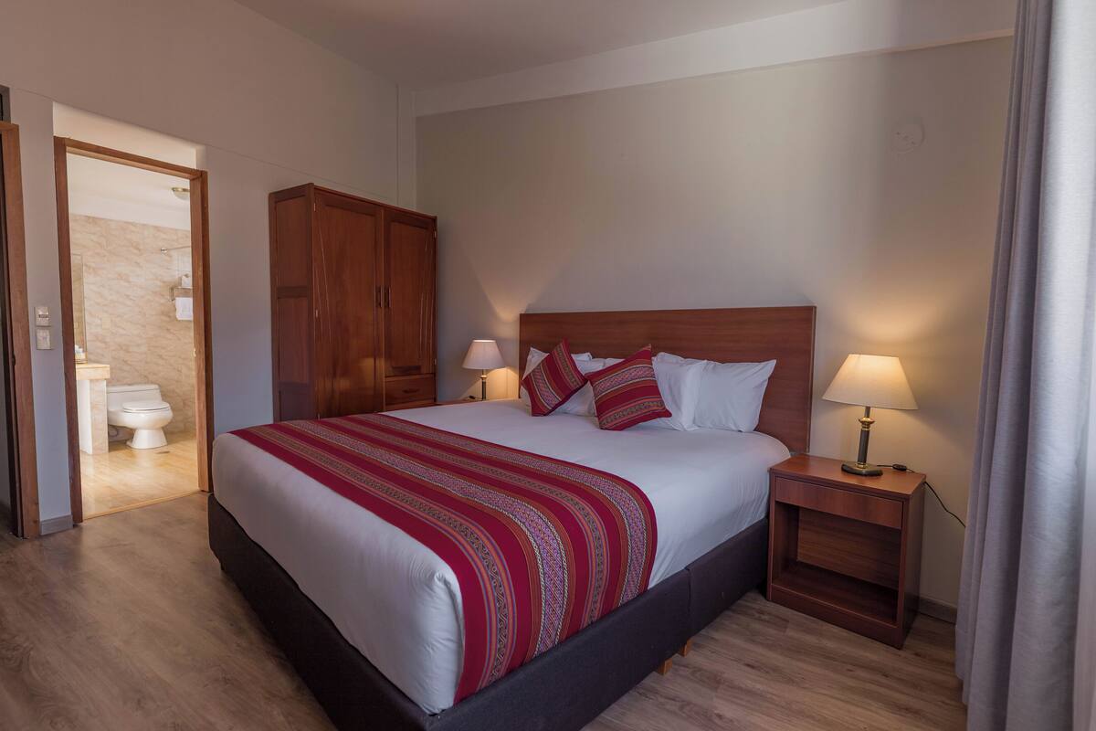 A hotel room with a large bed, wooden headboard, two bedside tables with lamps, a wardrobe, and a bathroom visible through an open door.