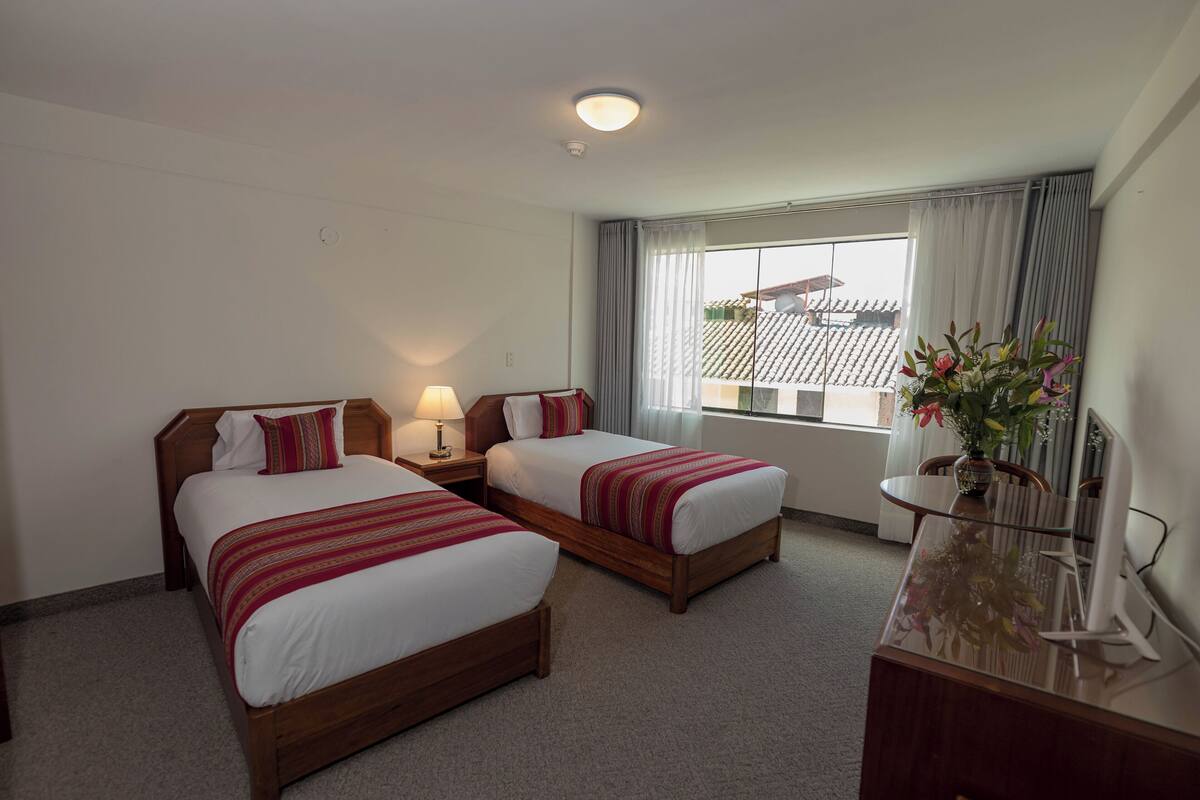A hotel room with two beds, a desk, a lamp, a vase with flowers, and a window with curtains.