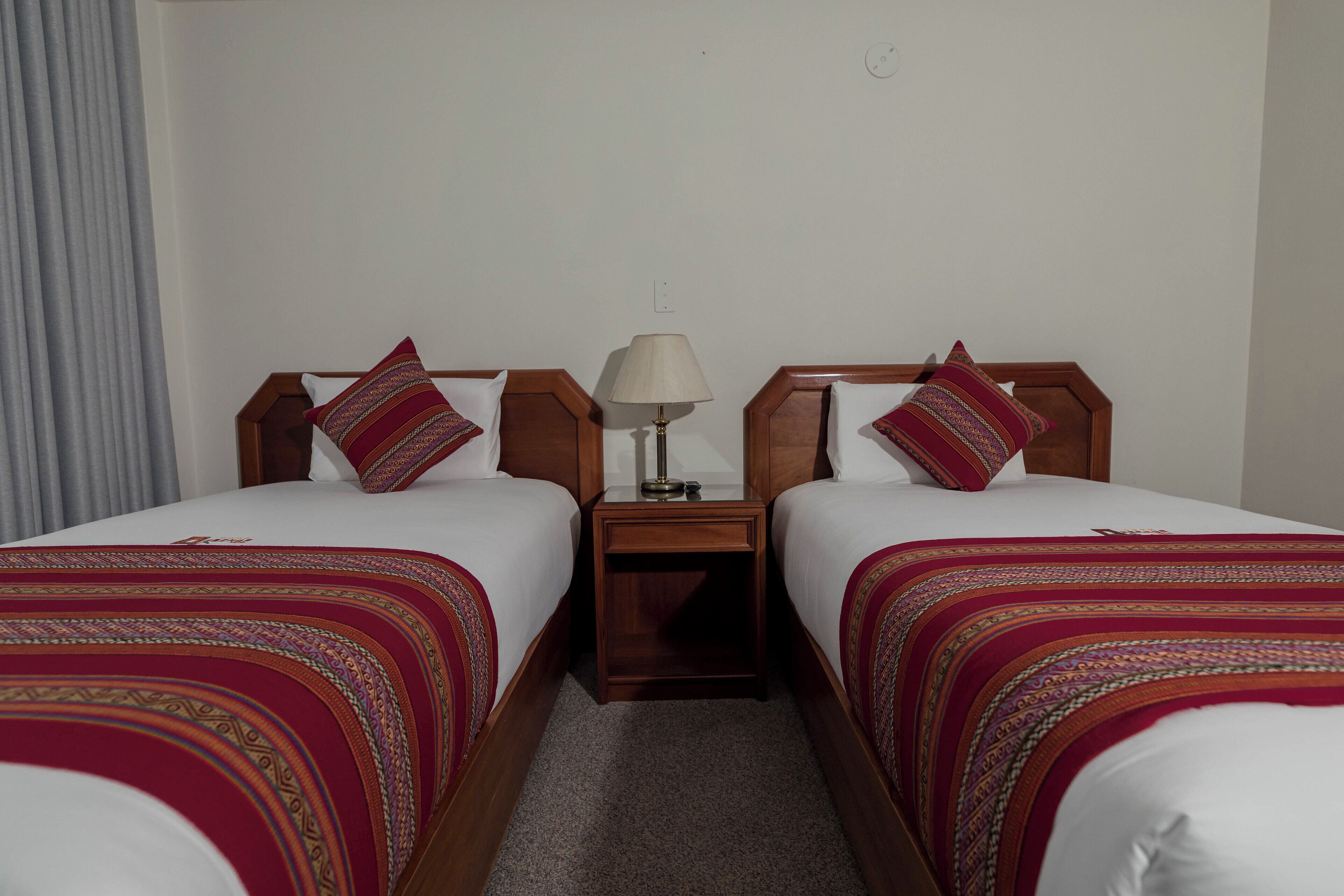 A hotel room with two single beds, each with a patterned bedspread and matching pillows. There is a nightstand with a lamp between the beds.