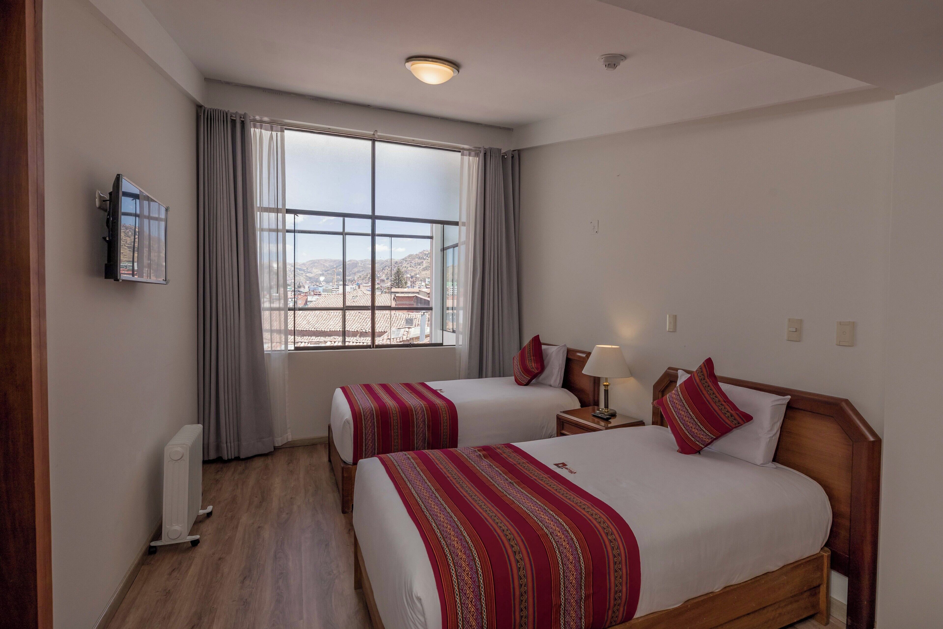 A hotel room with two beds, a window with curtains, a radiator, a TV on the wall, and a nightstand with a lamp.