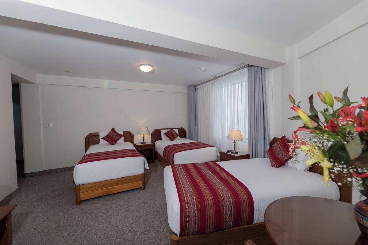 A hotel room with two beds, a desk, and a chair. There are lamps on the nightstands and a window with curtains.