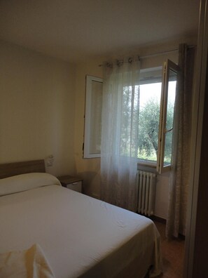 A bedroom with a bed, a nightstand, a window with curtains, and a radiator.