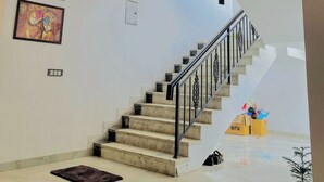 Un intérieur spacieux avec un sol en marbre, un escalier doté de rampes noires, un ventilateur de plafond et un tableau encadré accroché au mur.