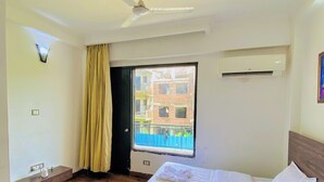 Une chambre d’hôtel avec un lit, un ventilateur de plafond, un appareil de climatisation, une fenêtre donnant sur des immeubles et une plante en pot.