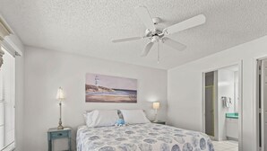 Un dormitorio con cama, ventilador de techo, pintura de un faro, mesita de noche con lámpara y baño visible a través de una puerta abierta.