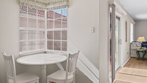 Una habitación con una mesa redonda blanca y dos sillas blancas, una ventana con cortinas estampadas y una puerta que da a otra estancia.
