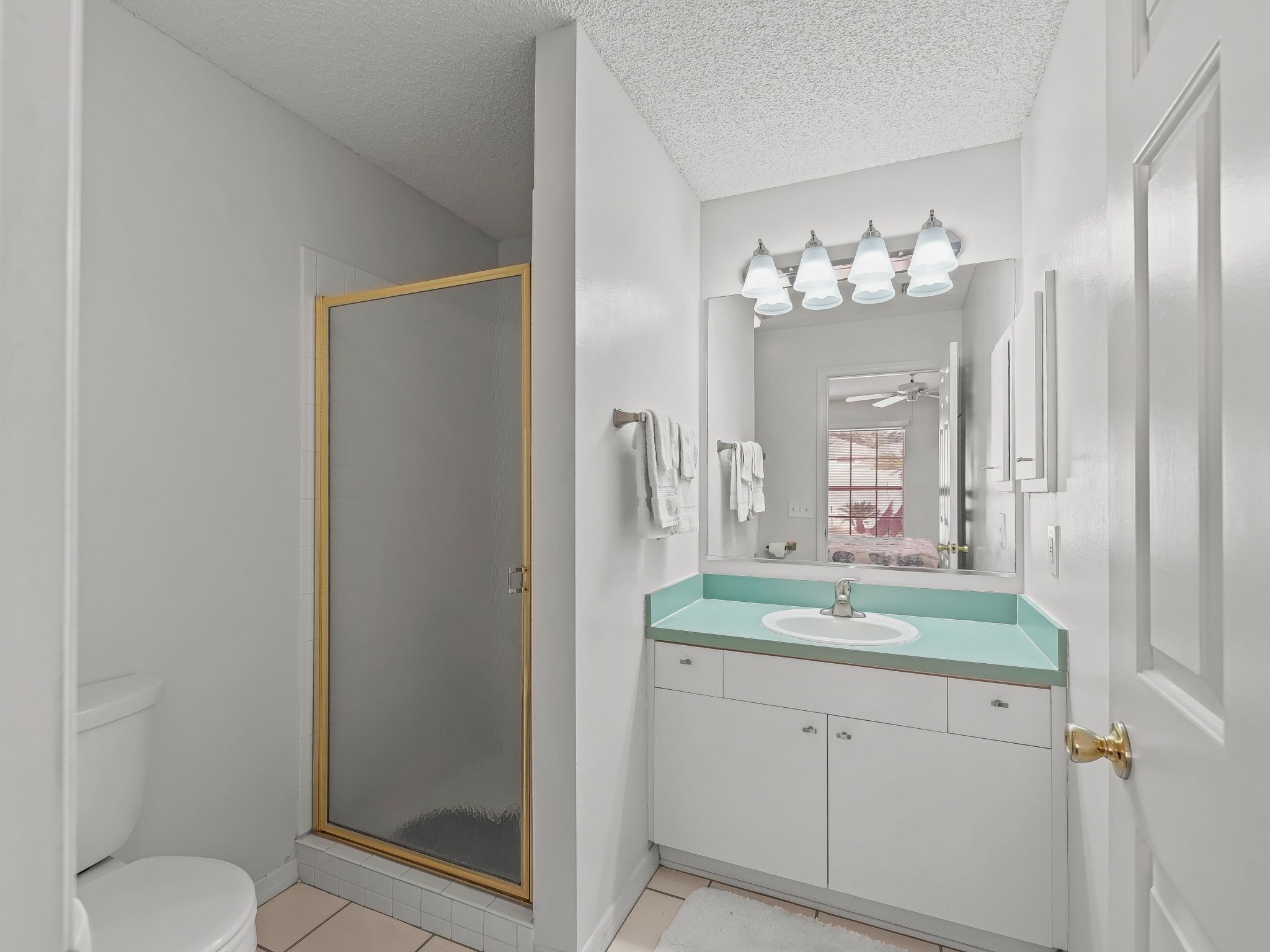 A bathroom with a white toilet, a glass shower enclosure, a sink with a turquoise countertop, and a mirror with three lights above it.