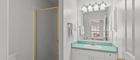 A bathroom with a white toilet, a glass shower enclosure, a sink with a turquoise countertop, and a mirror with three lights above it.