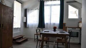 A dining area with a wooden table, chairs, and a bench. There is a window with curtains, a microwave, and a stove in the kitchen area. - Studio BEACH ENTRANCE and Free Parking (Ouistreham)