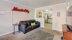 Ein Wohnzimmer mit grauem Sofa, einem kleinen Beistelltisch, einer TV-Konsole und einem Schild mit der Aufschrift 'Music City'.