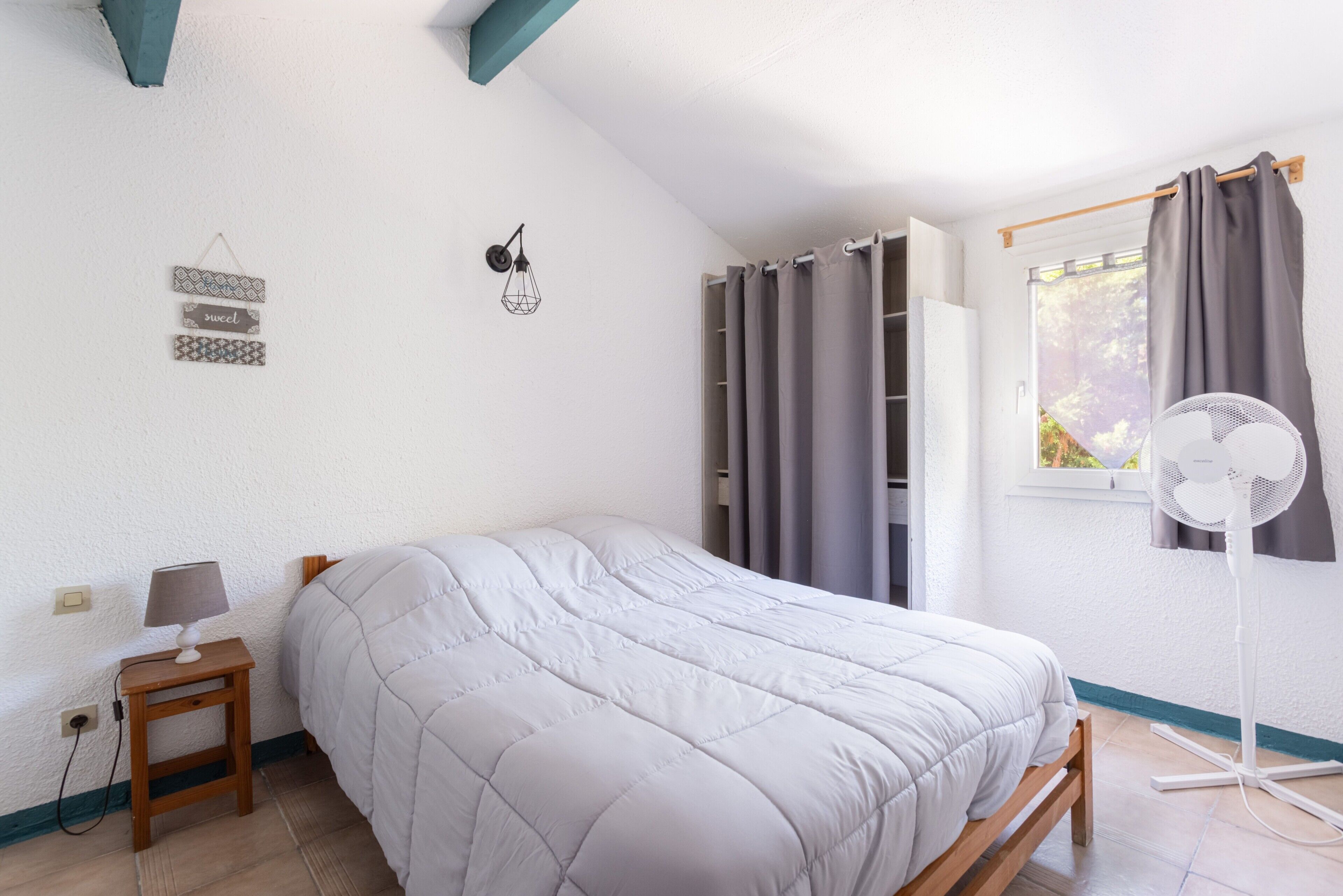Une chambre à coucher comprenant un lit, une table de chevet, une lampe, des éléments de décoration murale, des rideaux et un ventilateur sur pied.