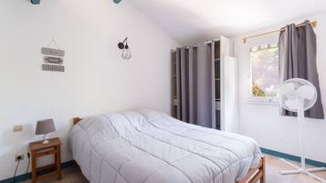 Une chambre à coucher comprenant un lit, une table de chevet, une lampe, des éléments de décoration murale, des rideaux et un ventilateur sur pied.