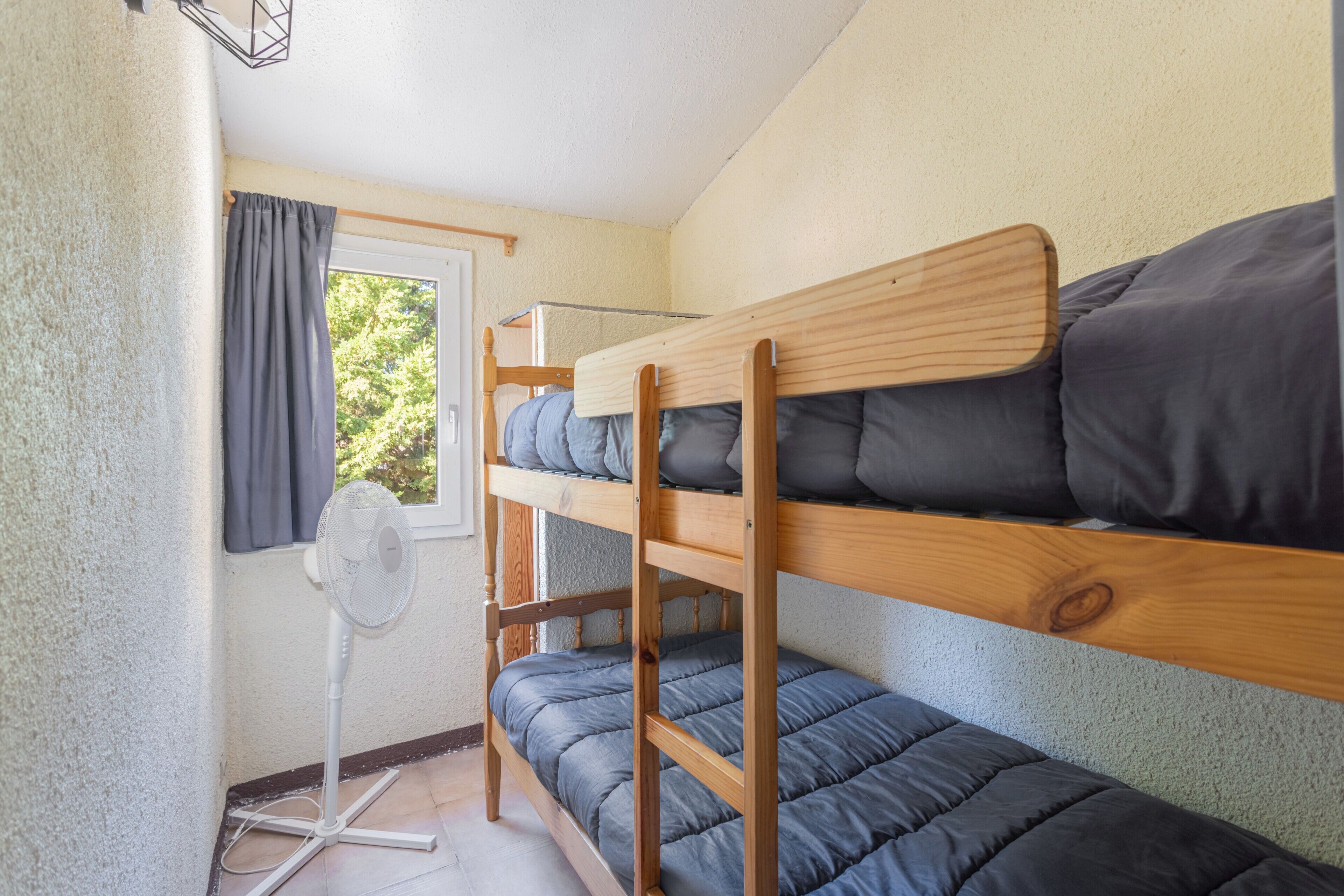 Une chambre avec un lit superposé, un ventilateur sur pied, une fenêtre avec des rideaux et une vue sur la verdure à l’extérieur.