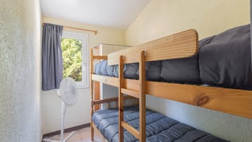Une chambre avec un lit superposé, un ventilateur sur pied, une fenêtre avec des rideaux et une vue sur la verdure à l’extérieur.