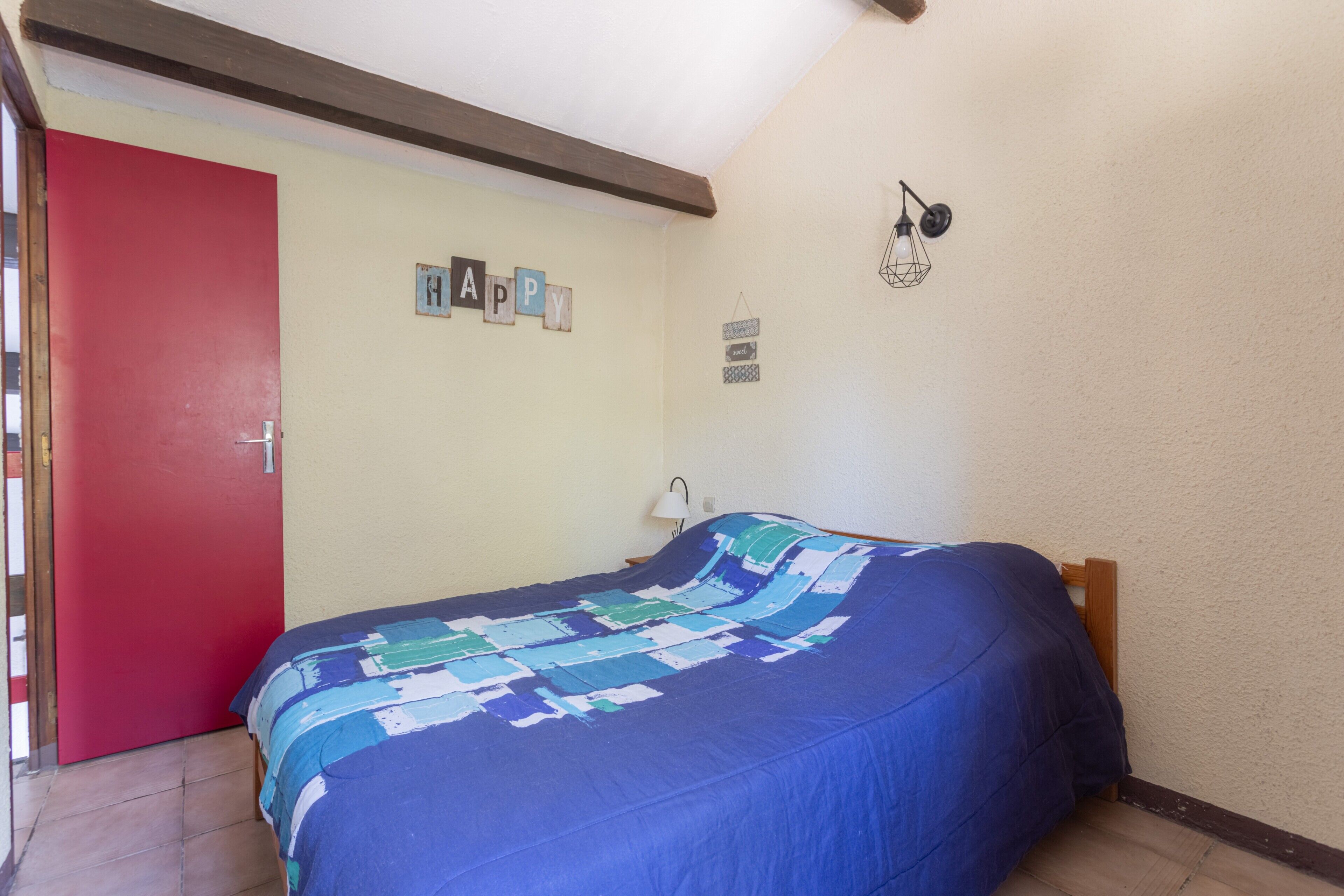 Una stanza con un letto, una porta rossa, un lampadario a parete e un cartello con la scritta 'HAPPY'.