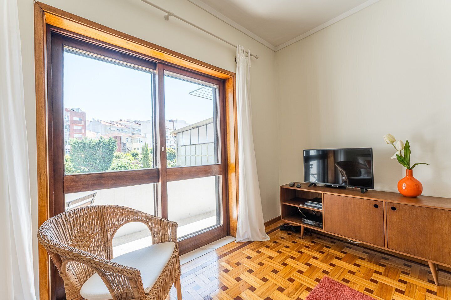 A room with a large sliding glass door, a wicker chair, a wooden TV stand with a flat-screen TV, and a red vase with flowers.