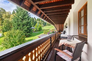 En balkon med gelænder af træ, et bord med stole, samt udsigt over et grønt landskab med træer og en græsplæne.