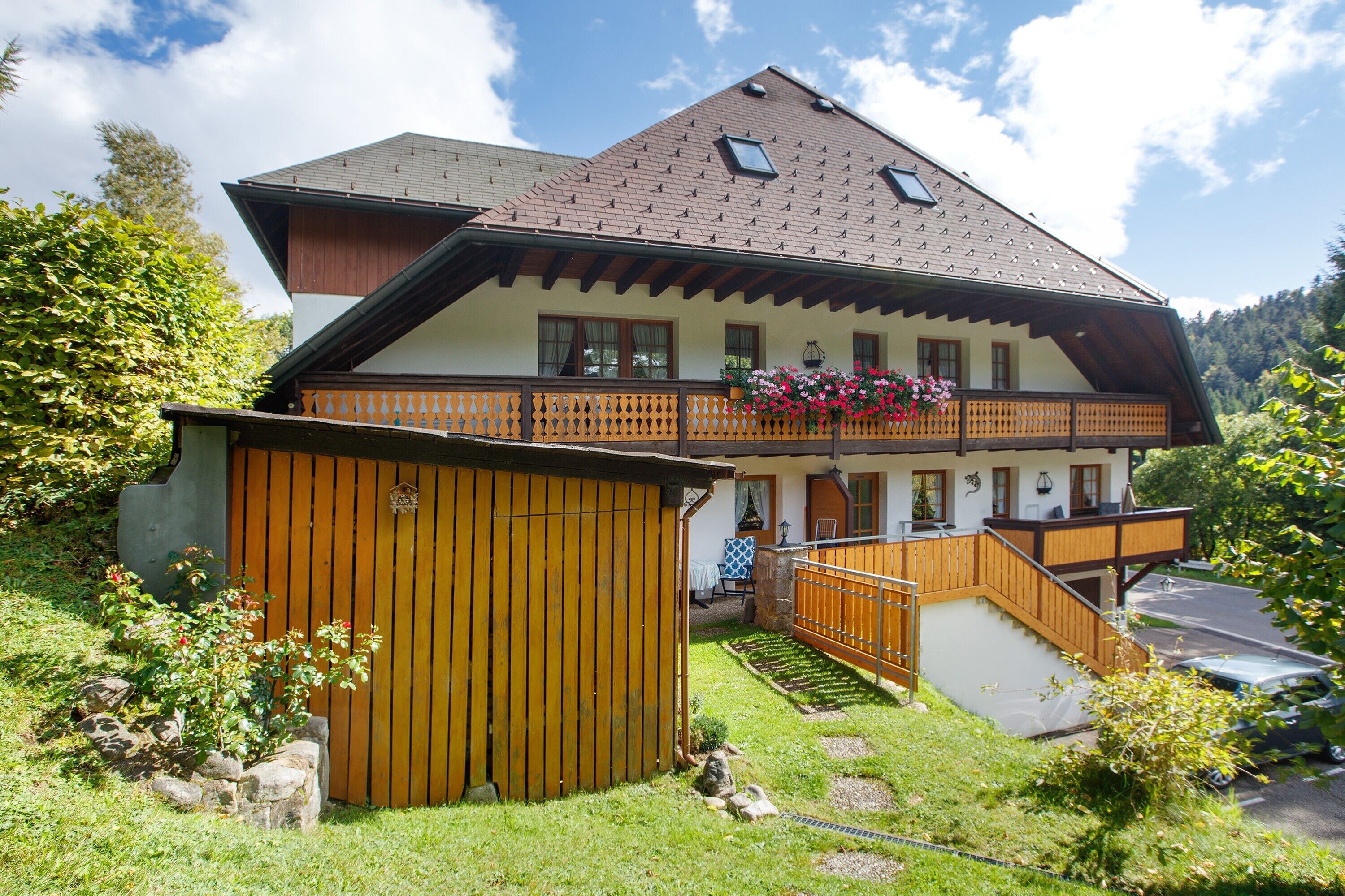 A two-story chalet with a wooden balcony, a stone wall, and a small garden.